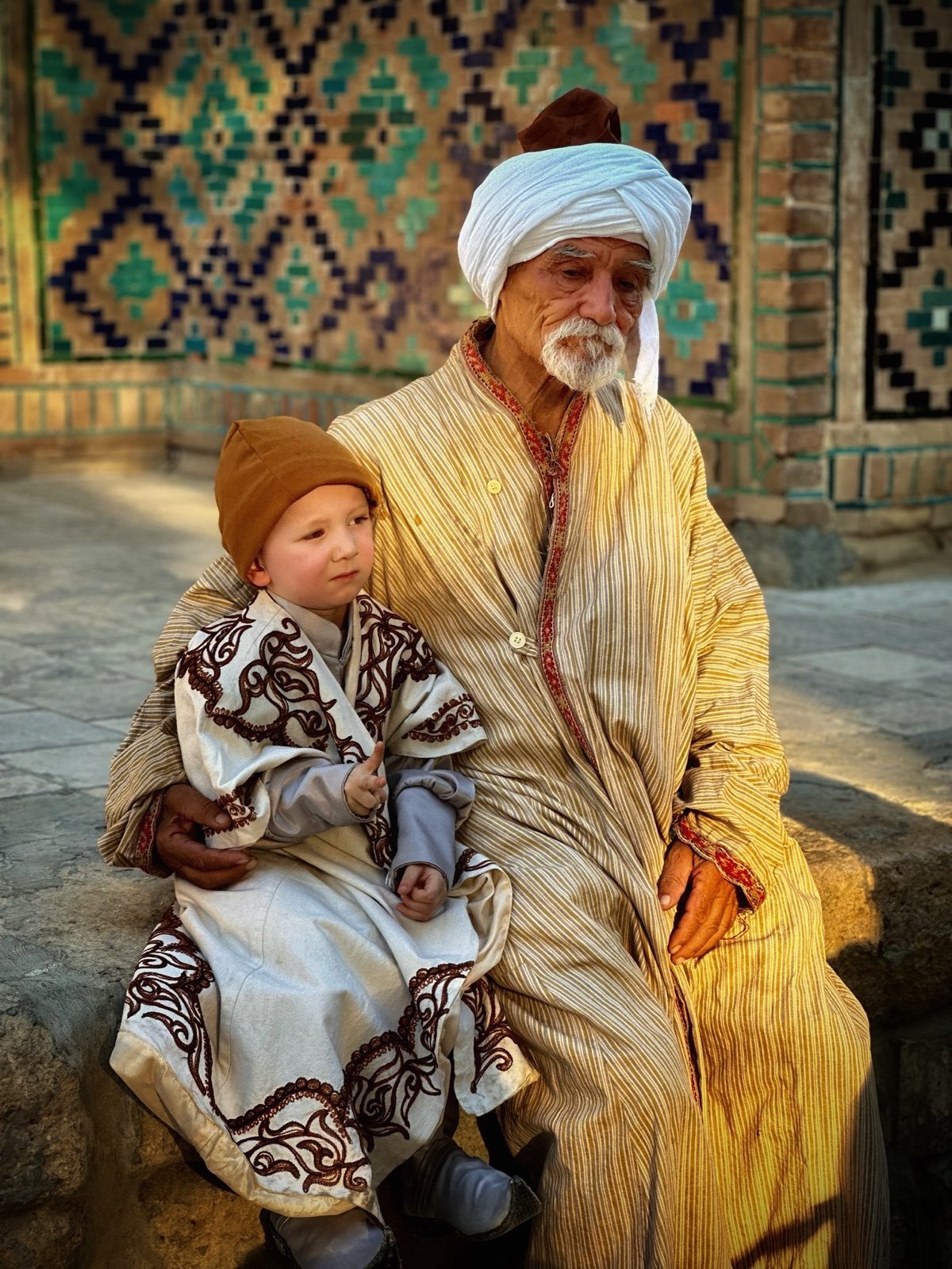 An older man and a child in traditional clothing beside patterned tilework in Bukhara