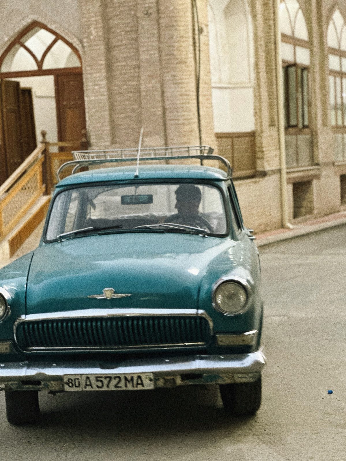 A turquoise vintage car parked beside old brick buildings in Bukhara